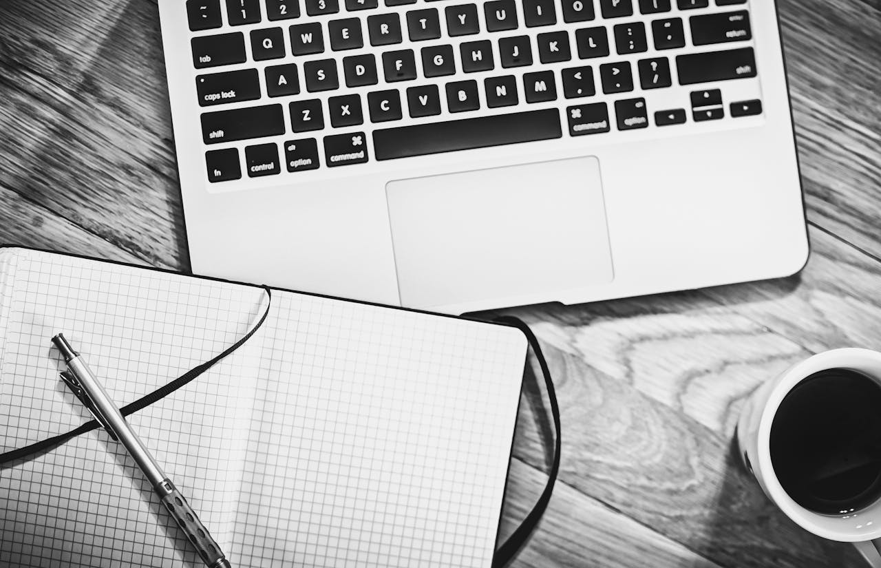 Home Black and white image of a workspace featuring a laptop, notebook, pen, and coffee cup.